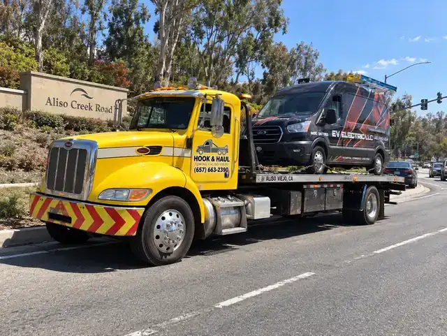 Yellow Hook & Haul flatbed tow truck carrying a van past the Aliso Creek Road sign in Aliso Viejo