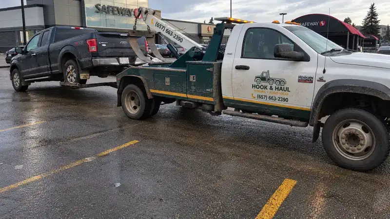 Green and white Hook & Haul tow truck towing a pickup in a wet parking lot